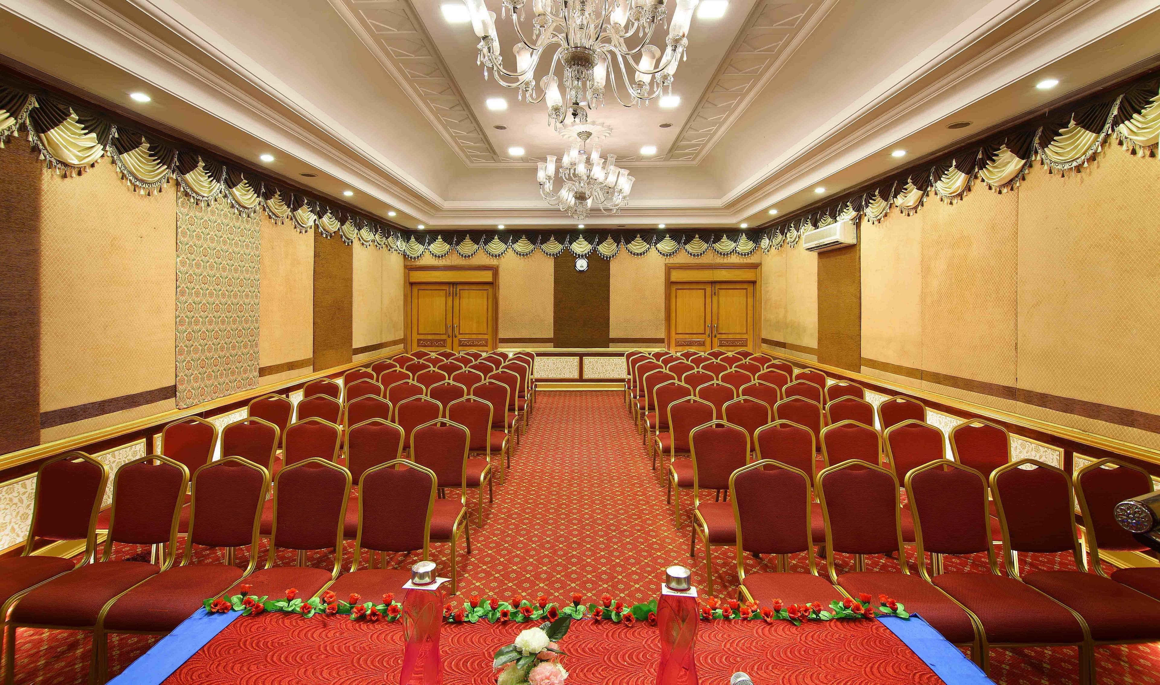 Un salón de conferencias con hileras de sillas rojas, una araña de cristal y paneles decorativos en las paredes.