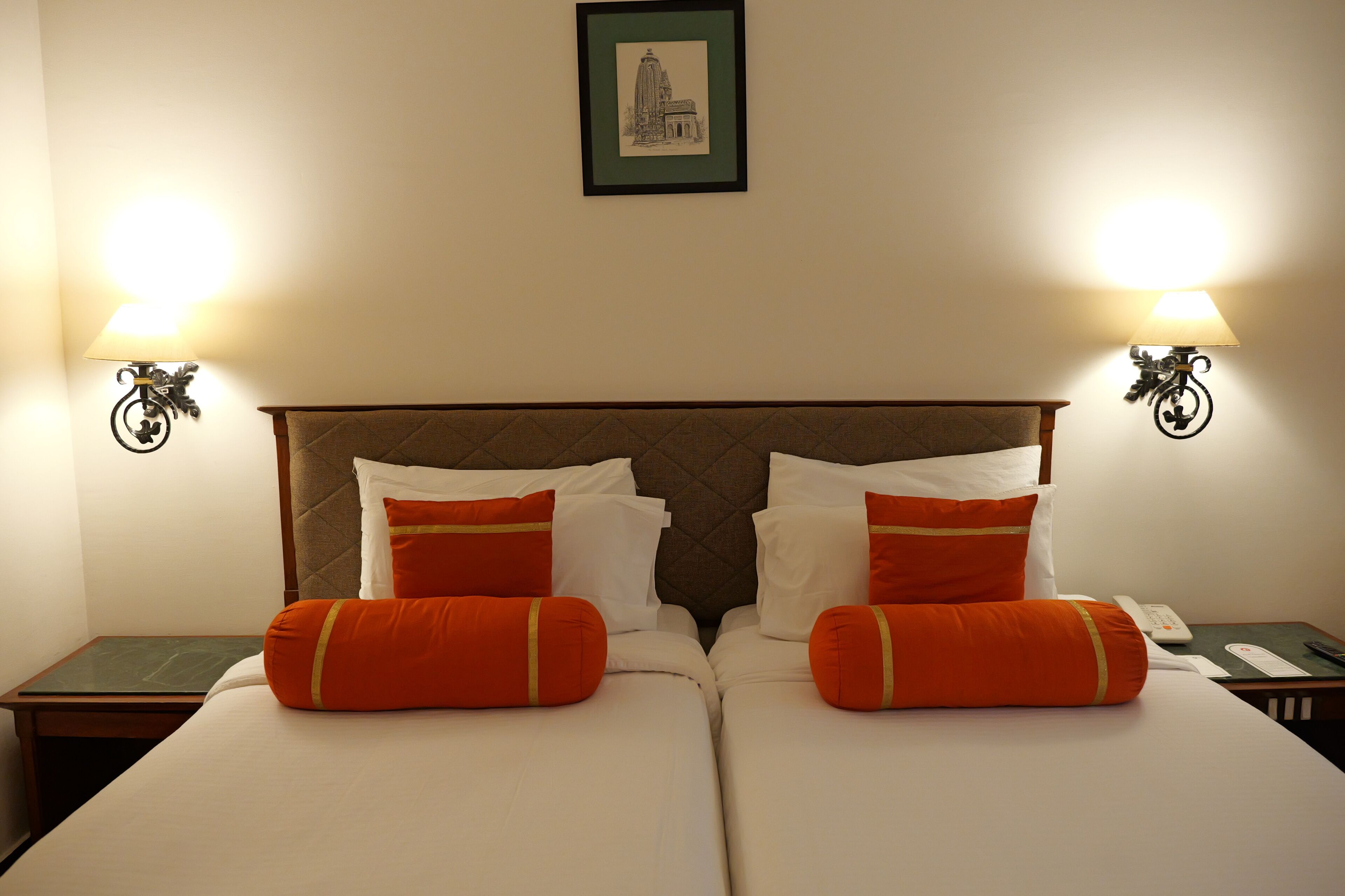 Una cama doble con ropa de cama blanca y naranja, dos mesitas de noche con lámparas y un cuadro en la pared.
