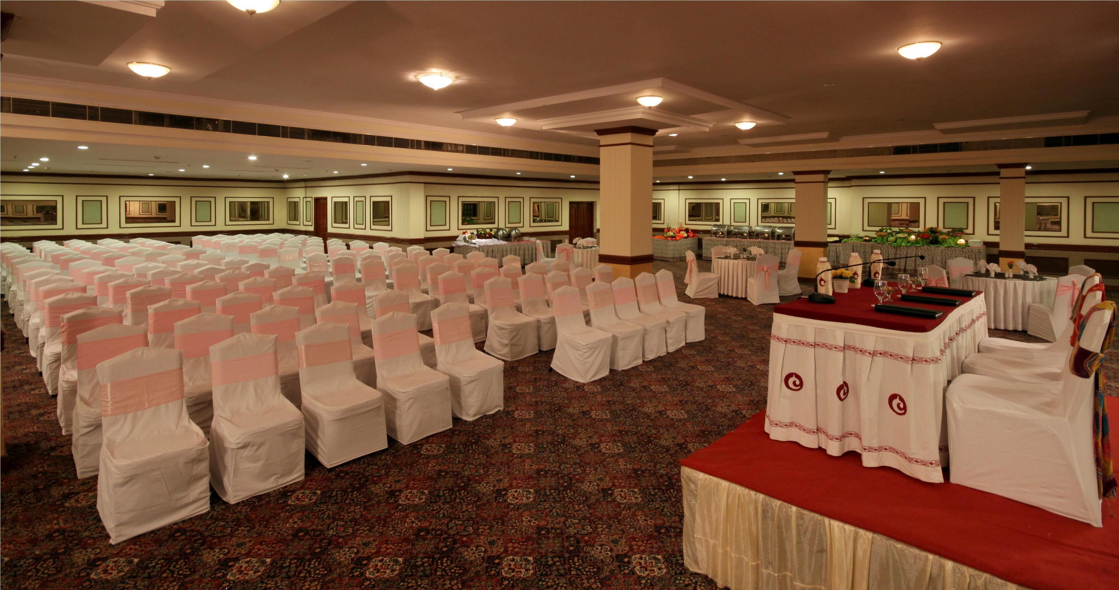 A large hall with rows of white chairs and tables, carpeted floor, and a stage area.