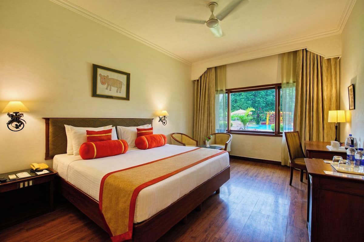 A hotel room with a large bed, two bedside lamps, a ceiling fan, a desk with a chair, and a window with curtains.