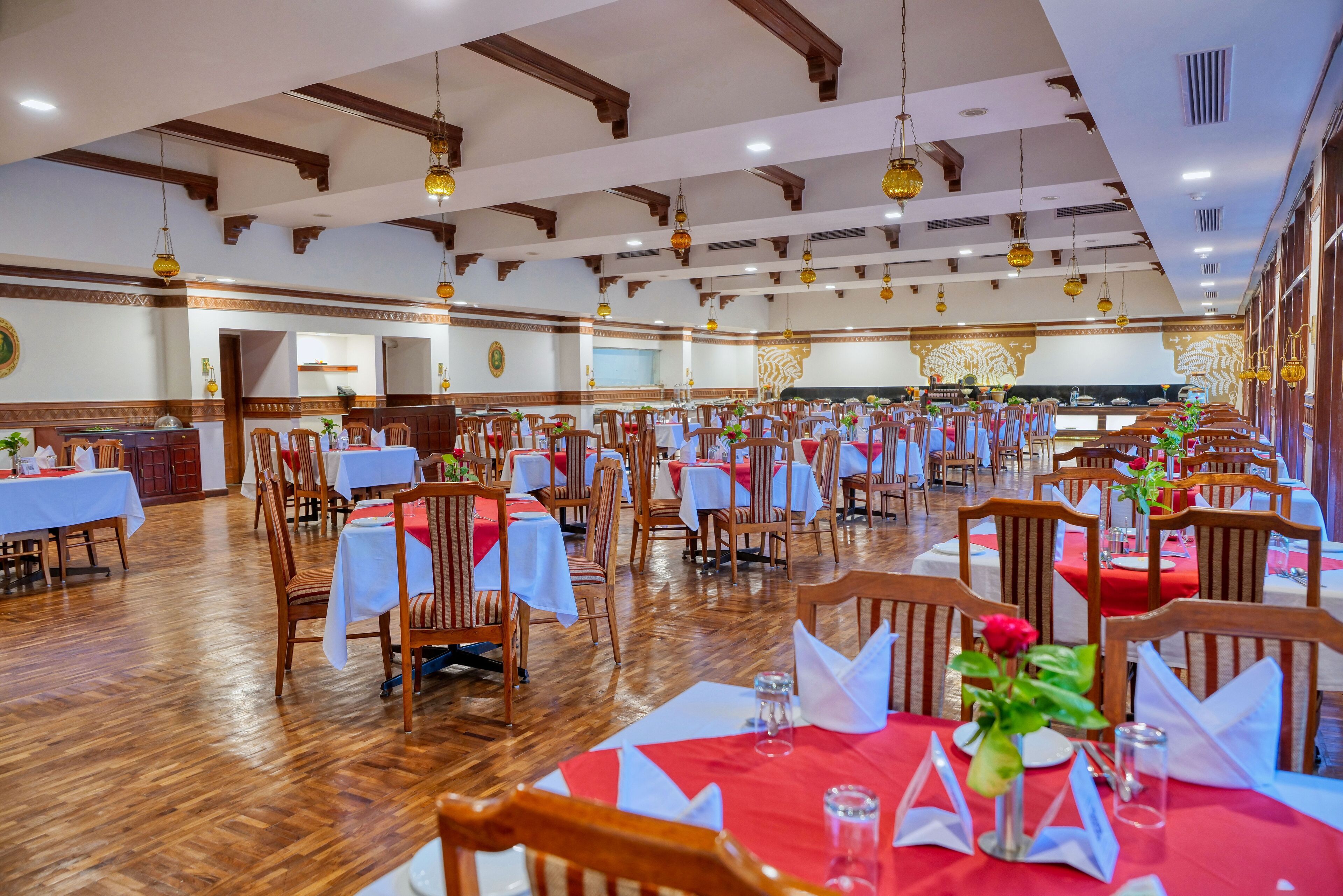 A large banquet hall with wooden floors and tables set for dining, featuring hanging light fixtures and a decorative wall.