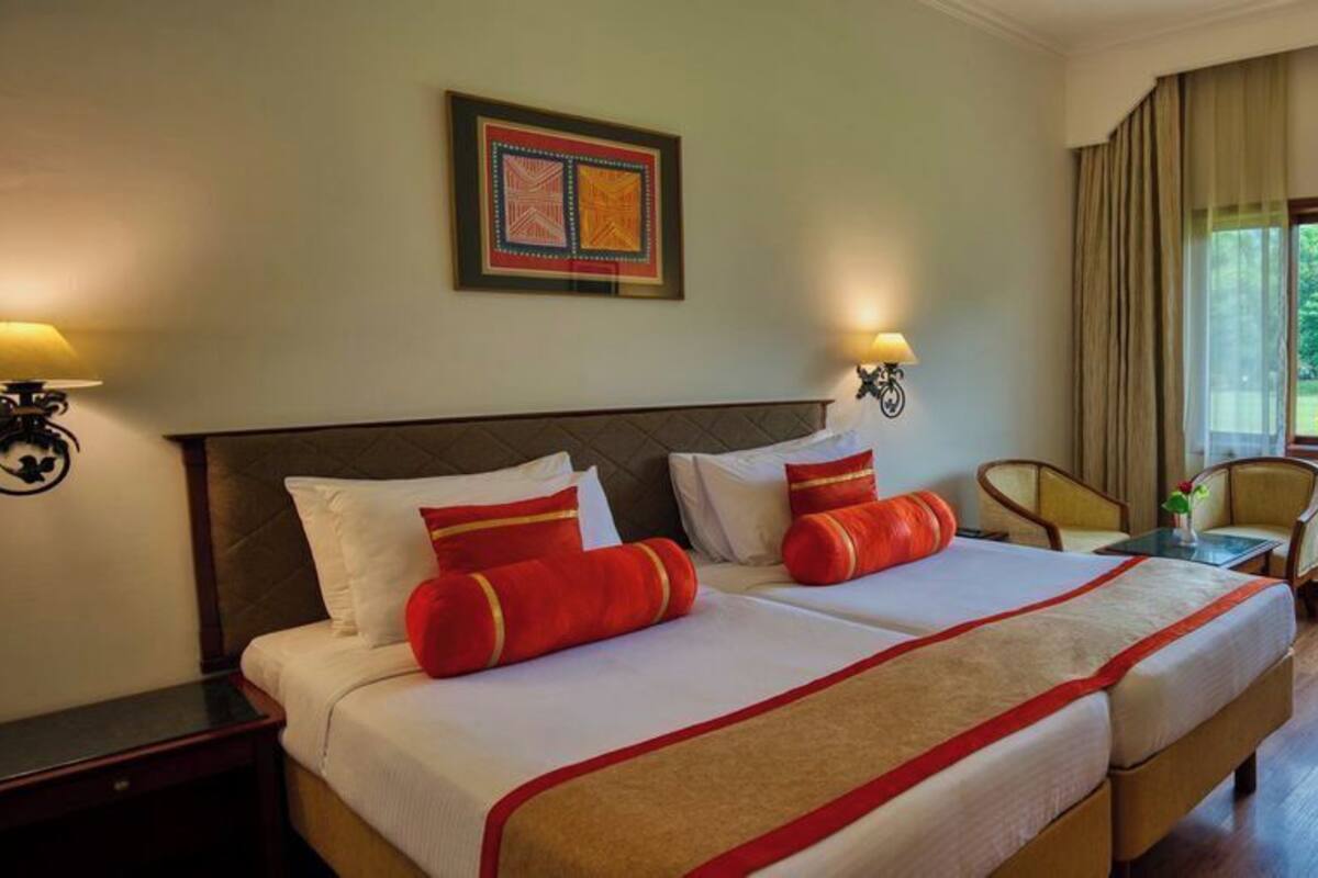A hotel room with two beds, a desk, a chair, and a window with a view of greenery.