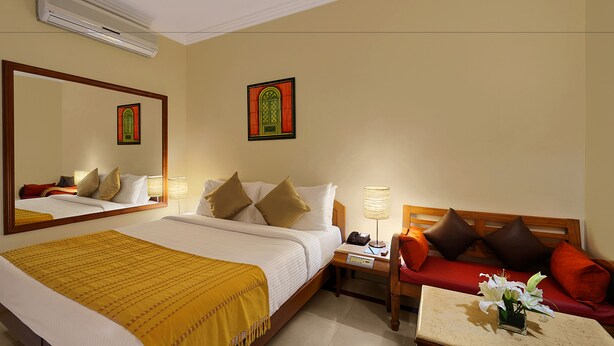 A hotel room with a bed, a bedside table with a lamp, a red sofa, and a small table with a vase of flowers. - Casa De Goa Boutique Resort (Calangute)
