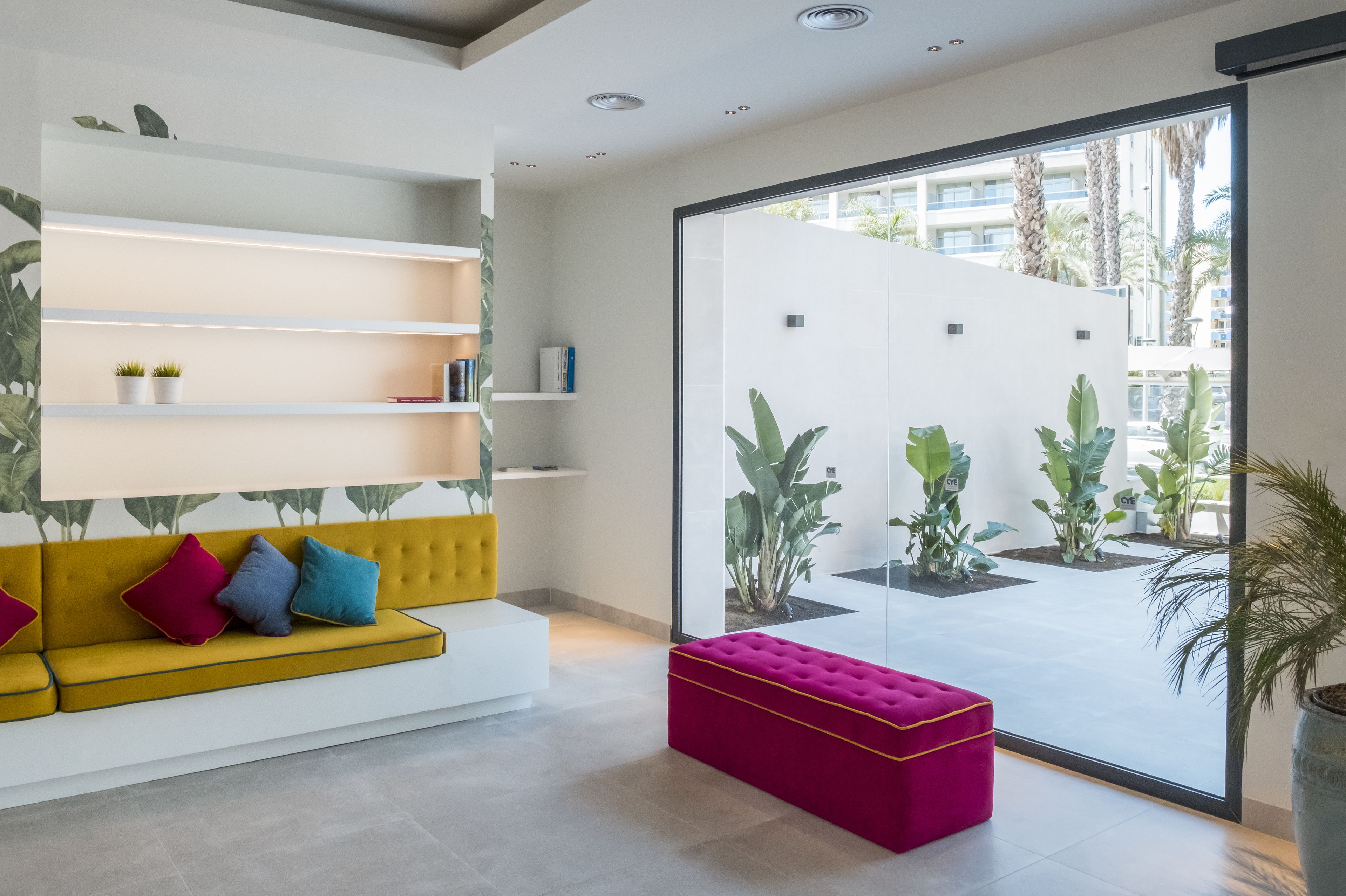 Un interior moderno con un sofá amarillo, un reposapiés rosa y una estantería blanca.