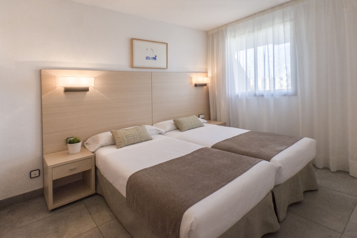 A hotel room with two beds, a nightstand, a wall-mounted light, a small potted plant, and a window with curtains.
