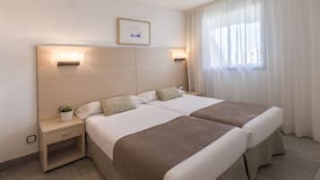 A hotel room with two beds, a nightstand, a wall-mounted light, a small potted plant, and a window with curtains.