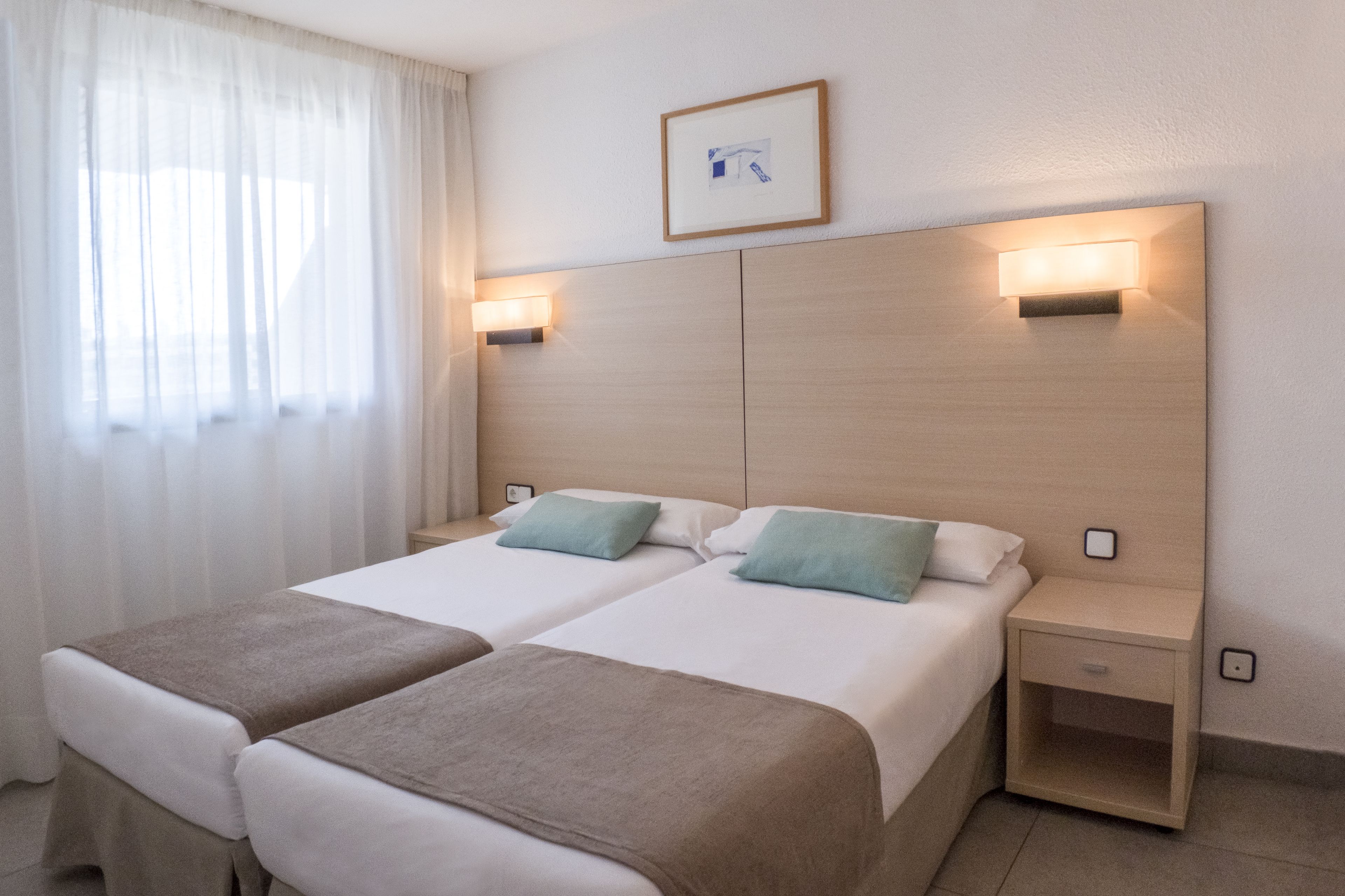 Ein Hotelzimmer mit zwei Betten, jedes mit einer braunen Decke und einem blauen Kissen. Es gibt Nachttische mit Lampen und ein gerahmtes Bild an der Wand.
