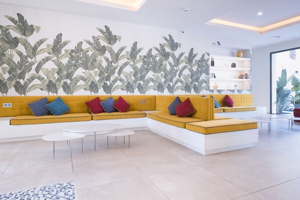 Un salon moderne avec un canapé d’angle jaune, des tables basses blanches et une fresque murale représentant des feuilles.