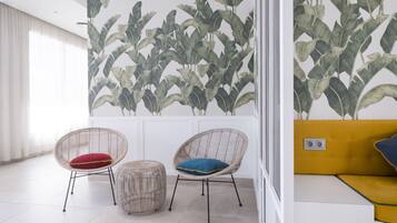 Una habitación con dos sillones de mimbre, uno con cojín rojo y el otro con cojín azul, un pequeño otoman redondo y un sofá amarillo con almohada azul.