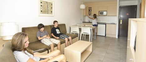A living room with a sofa, a coffee table, and a dining table with chairs. There is a kitchen area with a microwave and cabinets.