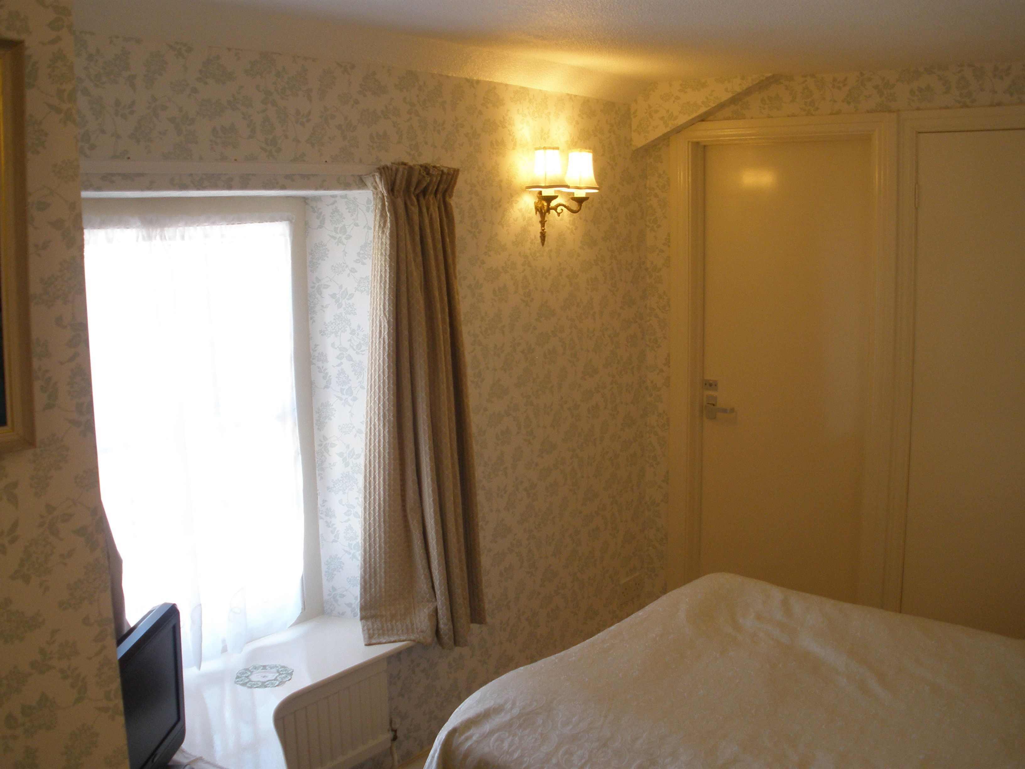 Une chambre d’hôtel équipée d’un lit, d’un bureau, d’une fenêtre avec des rideaux et d’un luminaire fixé au mur.