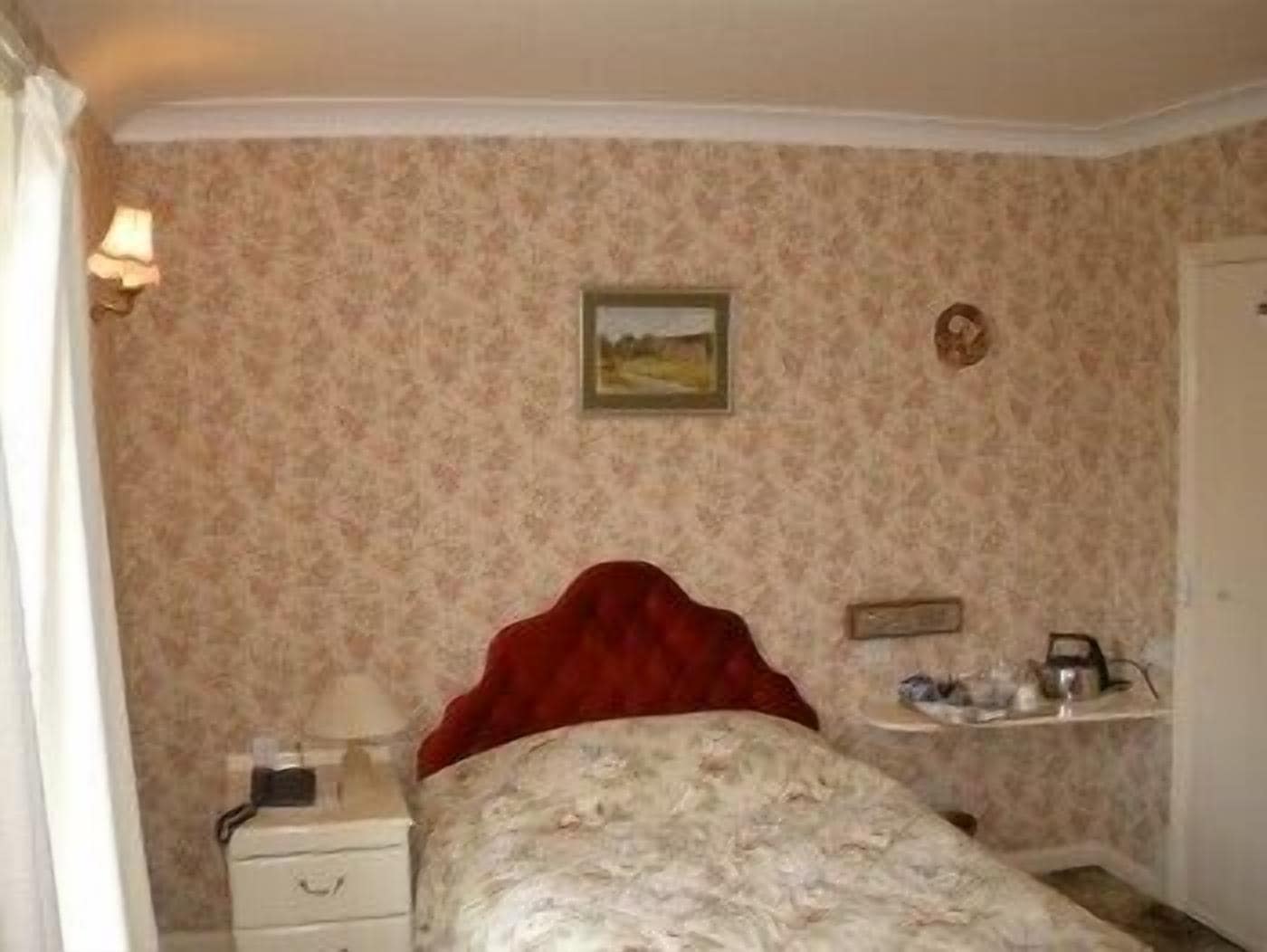 Une chambre à coucher avec un papier peint à motifs floraux, une tête de lit rouge, une table de chevet avec une lampe et une étagère contenant des articles de toilette.
