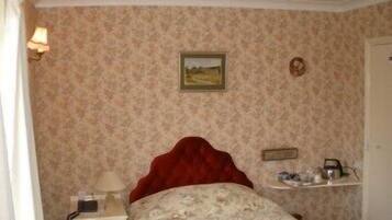 Une chambre à coucher avec un papier peint à motifs floraux, une tête de lit rouge, une table de chevet avec une lampe et une étagère contenant des articles de toilette.