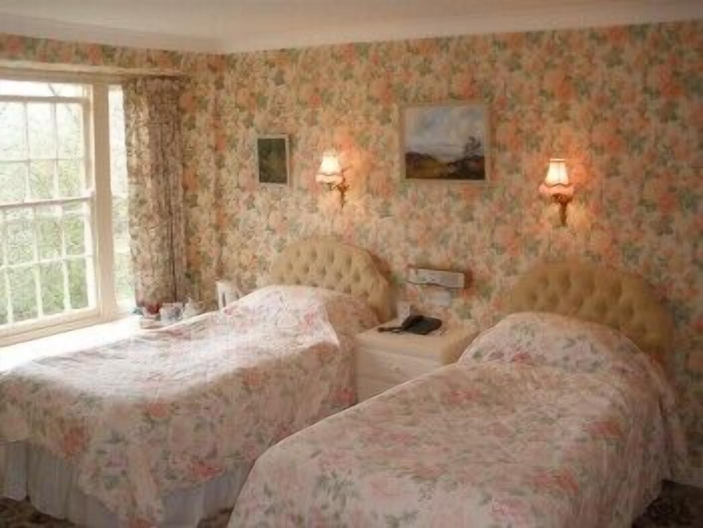 A room with two beds, floral wallpaper, and a window.