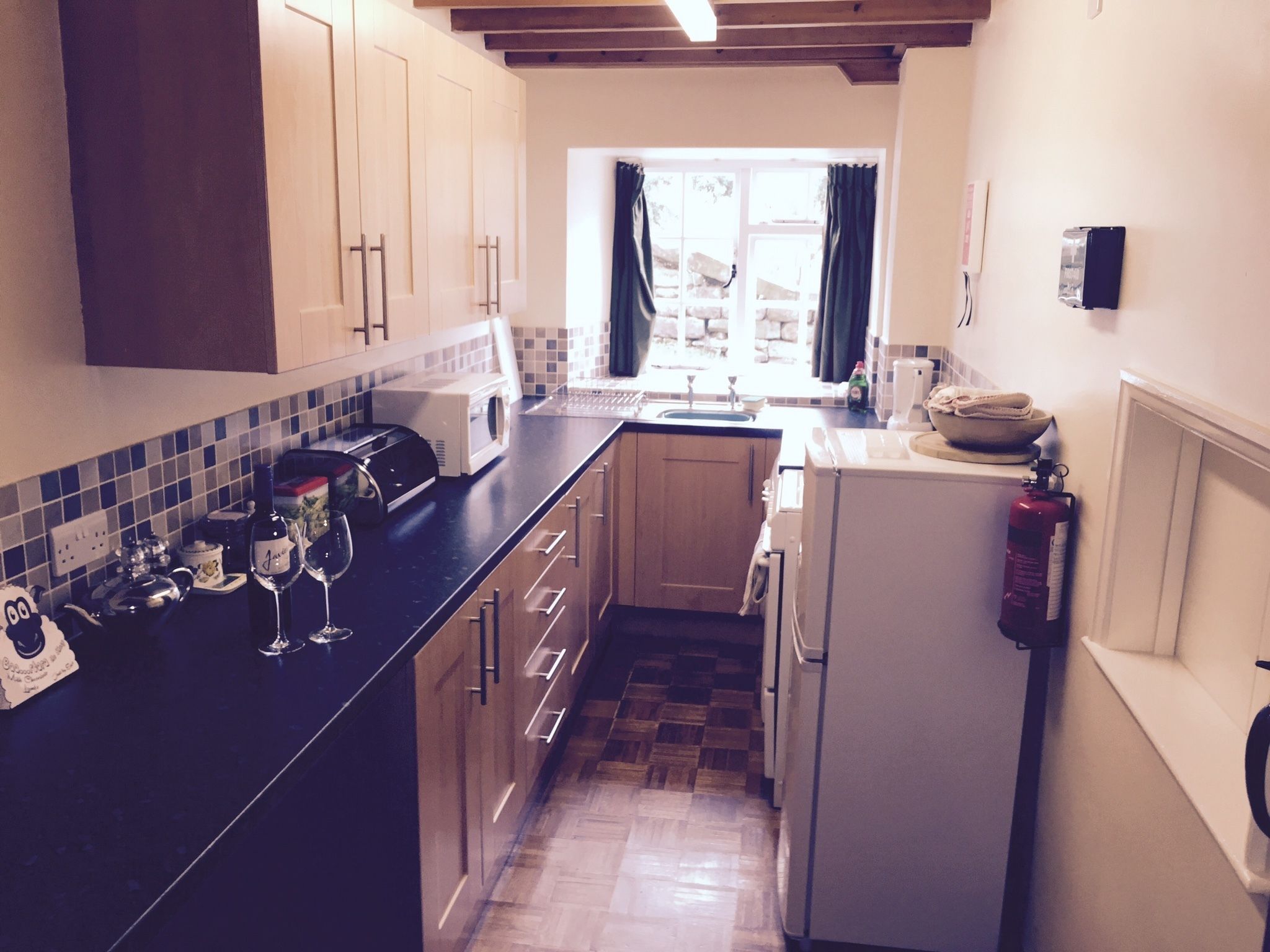 A compact kitchen with wooden cabinets, a black countertop, a white refrigerator, and a window with curtains.
