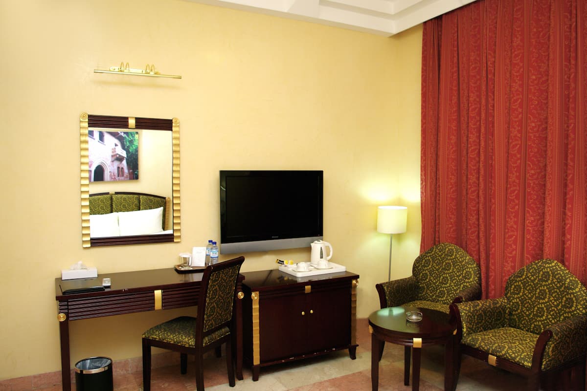 Habitación de hotel con televisión, escritorio, dos sillas, una mesita, lámpara y cubo de basura.
