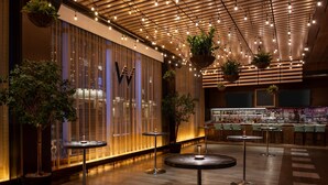 Uma área de bar moderna com mesas redondas, plantas pendentes e teto de madeira com iluminação.