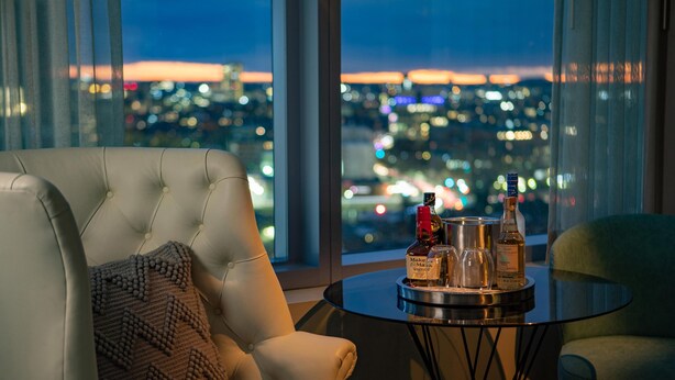 Quarto com vista para a paisagem urbana noturna, com uma poltrona de couro, uma mesa redonda pequena com garrafas e outra poltrona verde.