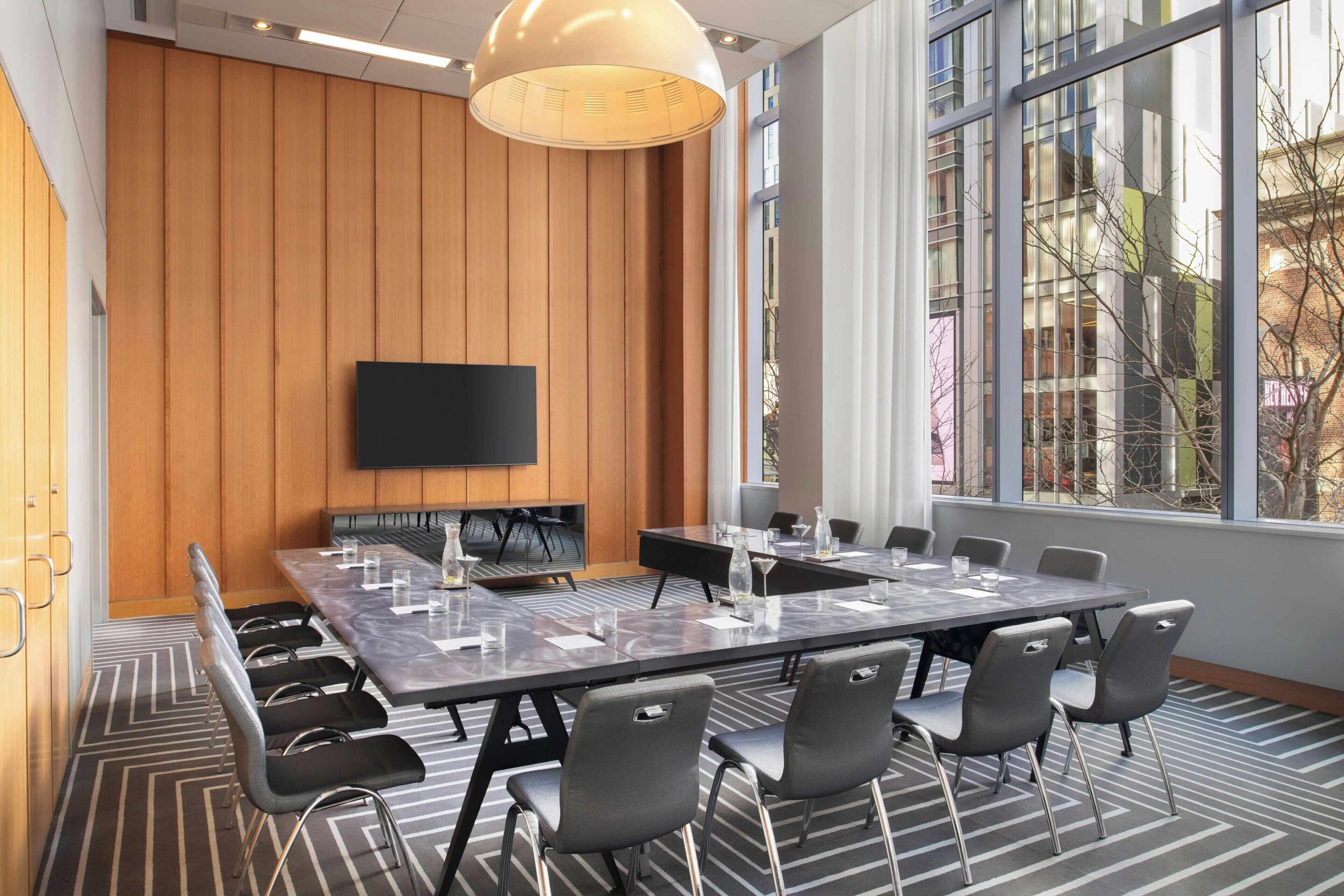 Un salón de conferencias con una gran mesa, sillas, un televisor de pantalla plana y un ventanal con vistas a edificios.
