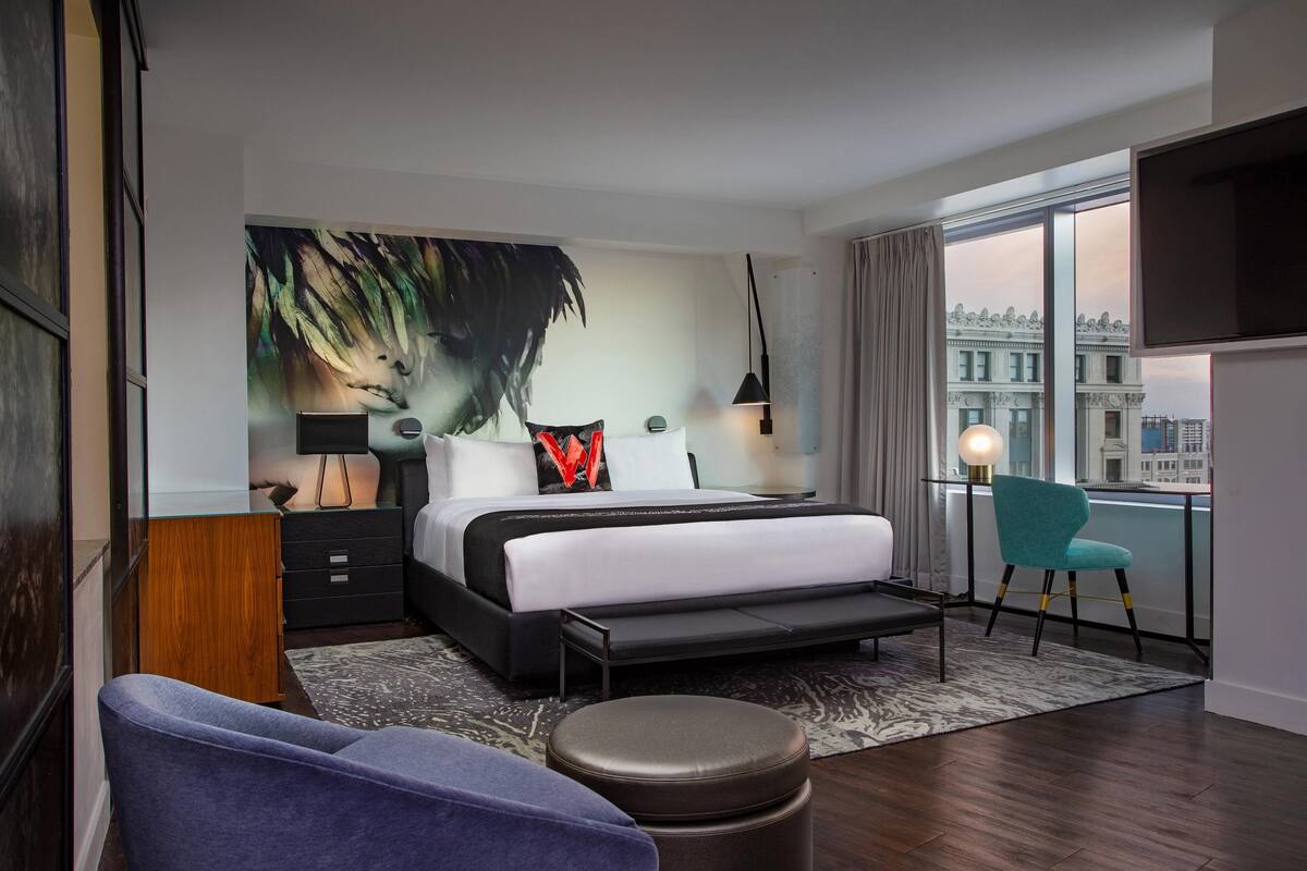 Een moderne hotelkamer met een groot bed, een bureau, een stoel en uitzicht op de stad door grote ramen.
