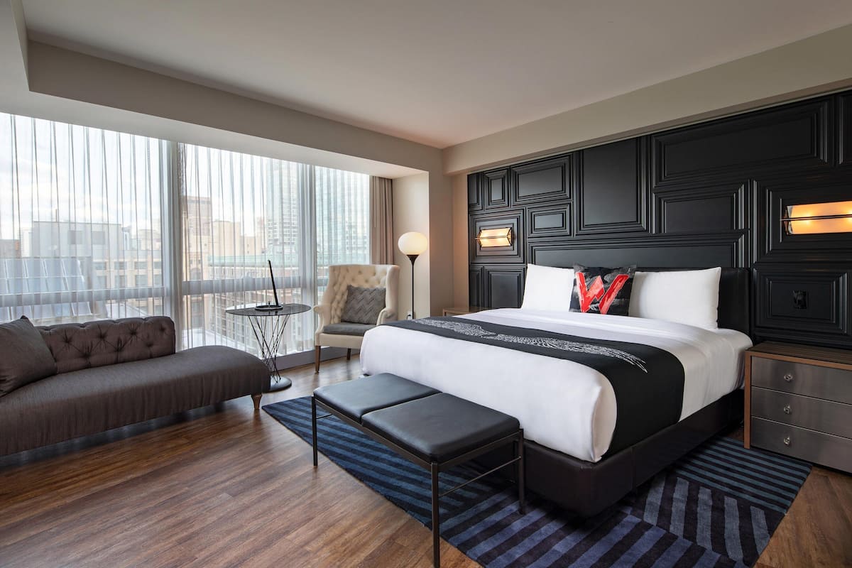 Een moderne hotelkamer met een groot bed, een bank, een zitbankje en uitzicht op de stad door grote ramen.