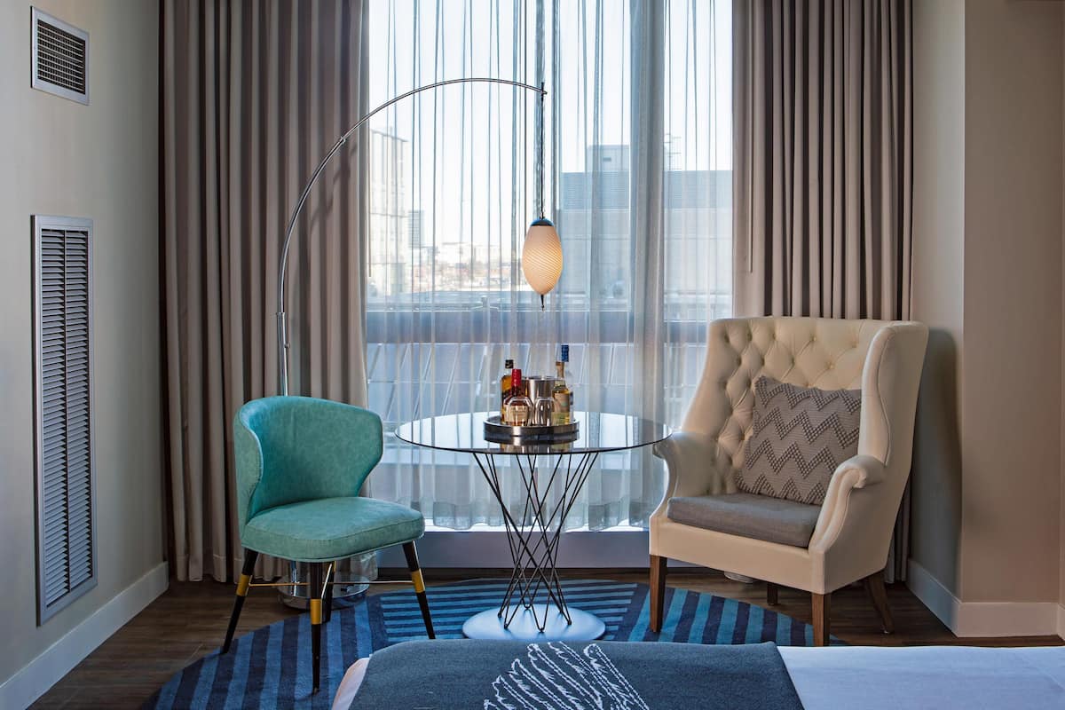 Een hotelkamer met een groot raam, een gestofte fauteuil, een smaragdgroene fauteuil, een glazen tafel met flessen en een blauw tapijt met patroon.