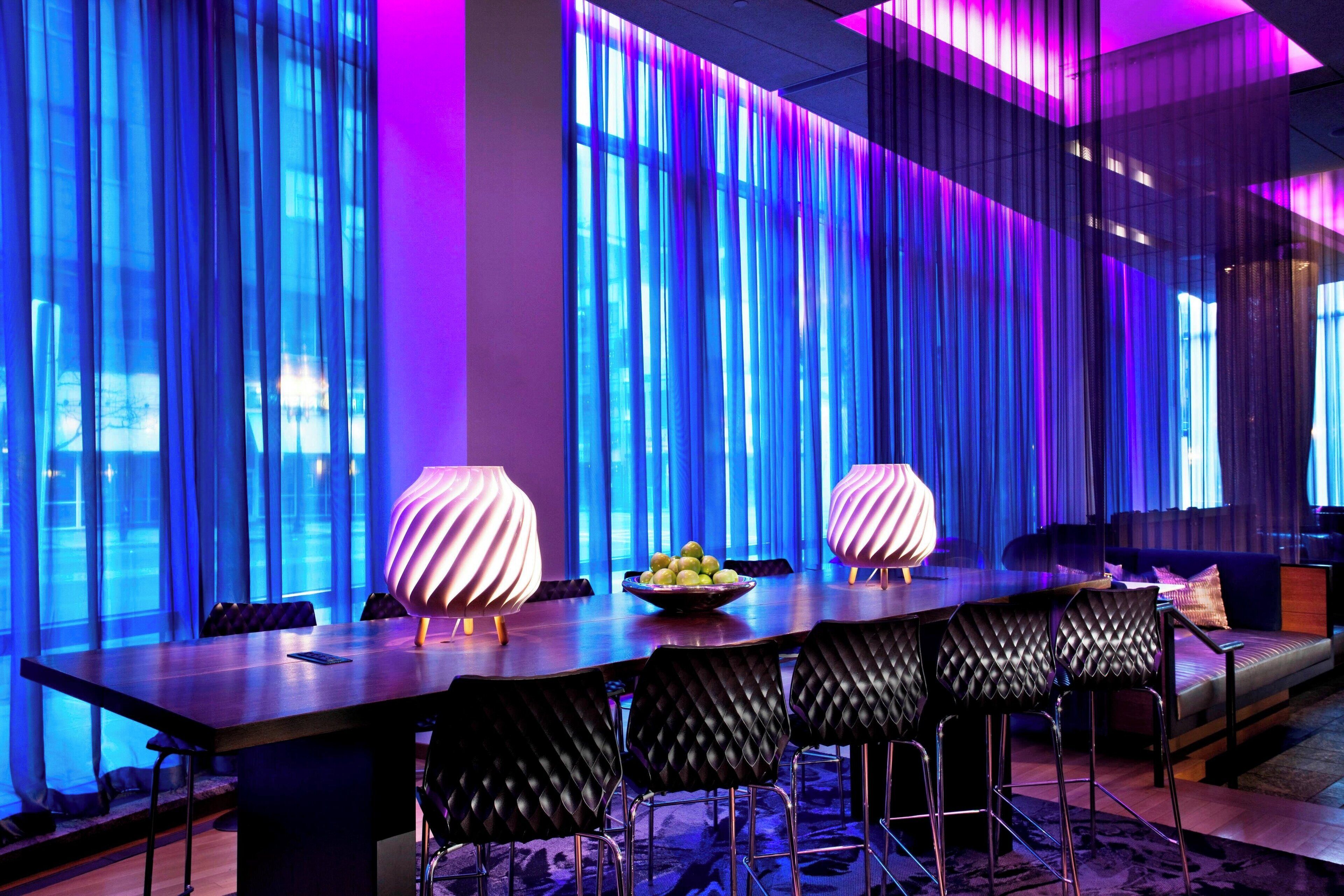 Un comedor moderno con una mesa larga, sillas negras acolchadas y dos lámparas iluminadas.