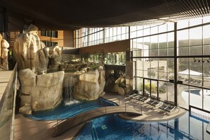 Une piscine intérieure agrémentée d’une cascade, de chaises longues et de grandes fenêtres offrant une vue sur les environs.