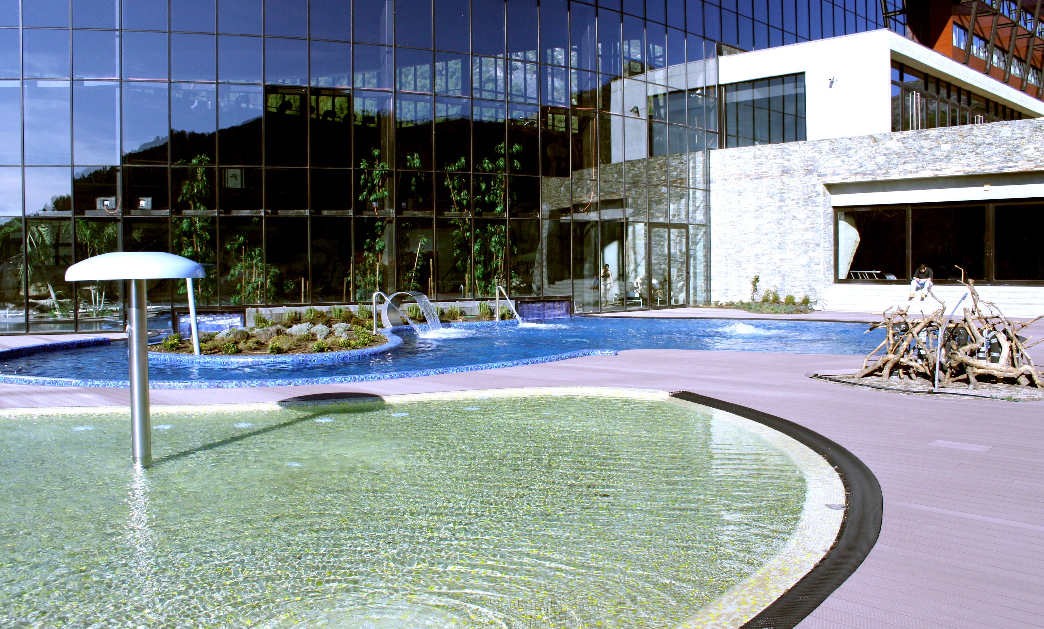 Un edificio moderno con fachada de vidrio, una piscina con fuente de agua y un muro de piedra.