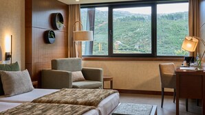 A hotel room with a large bed, two armchairs, a desk, and a view of a green landscape through a large window. - H2otel Congress & Medical Spa (Covilhã)