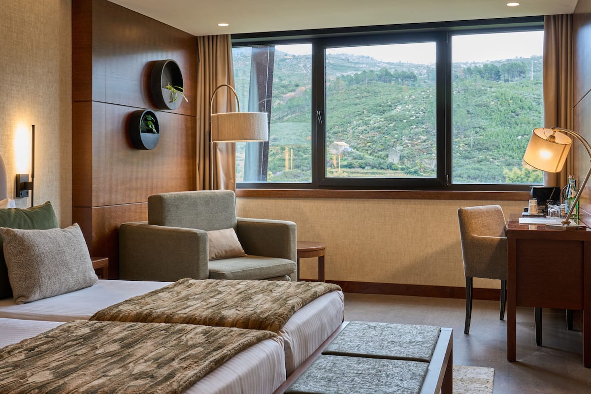 A hotel room with a large bed, two armchairs, a desk, and a view of a green landscape through a large window.