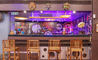 Zona de bar con sillas de madera, un cartel de salida de incendios y máscaras de colores en la pared.