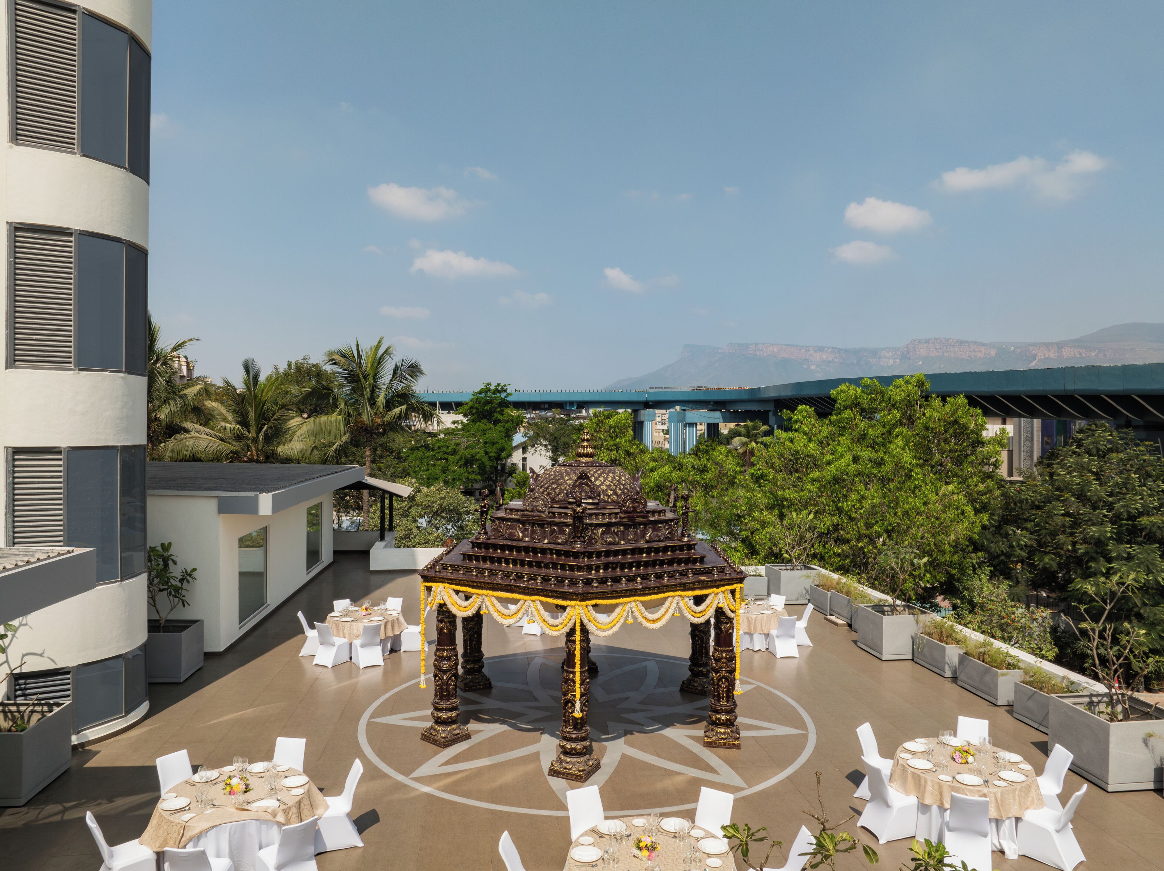 An outdoor dining area with a central gazebo, surrounded by white tables and chairs, set against a backdrop of a modern building and a distant mountain range.