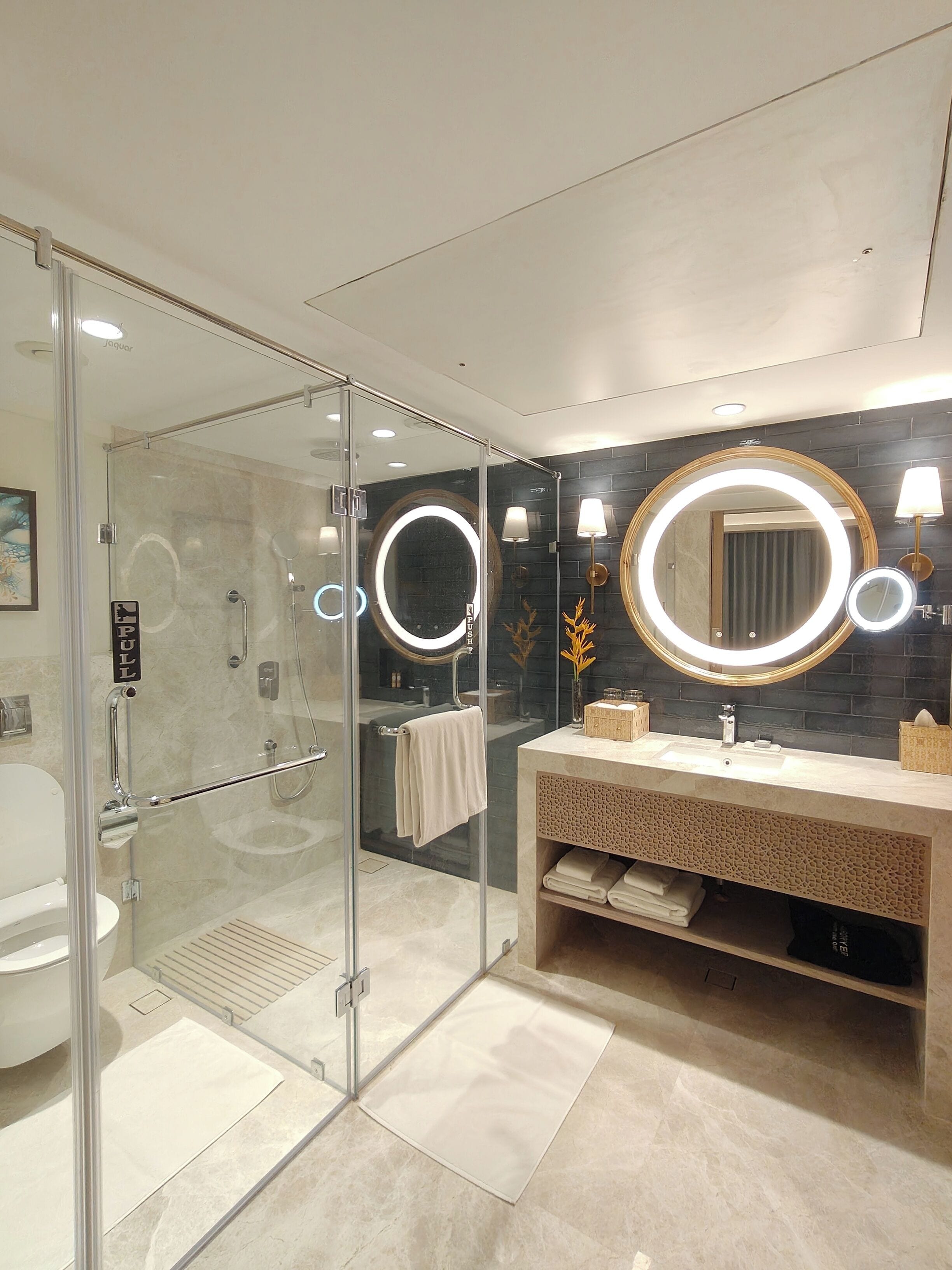 Un baño moderno con ducha envuelta en vidrio, tocador con espejo redondo y salpicadero de azulejos oscuros.