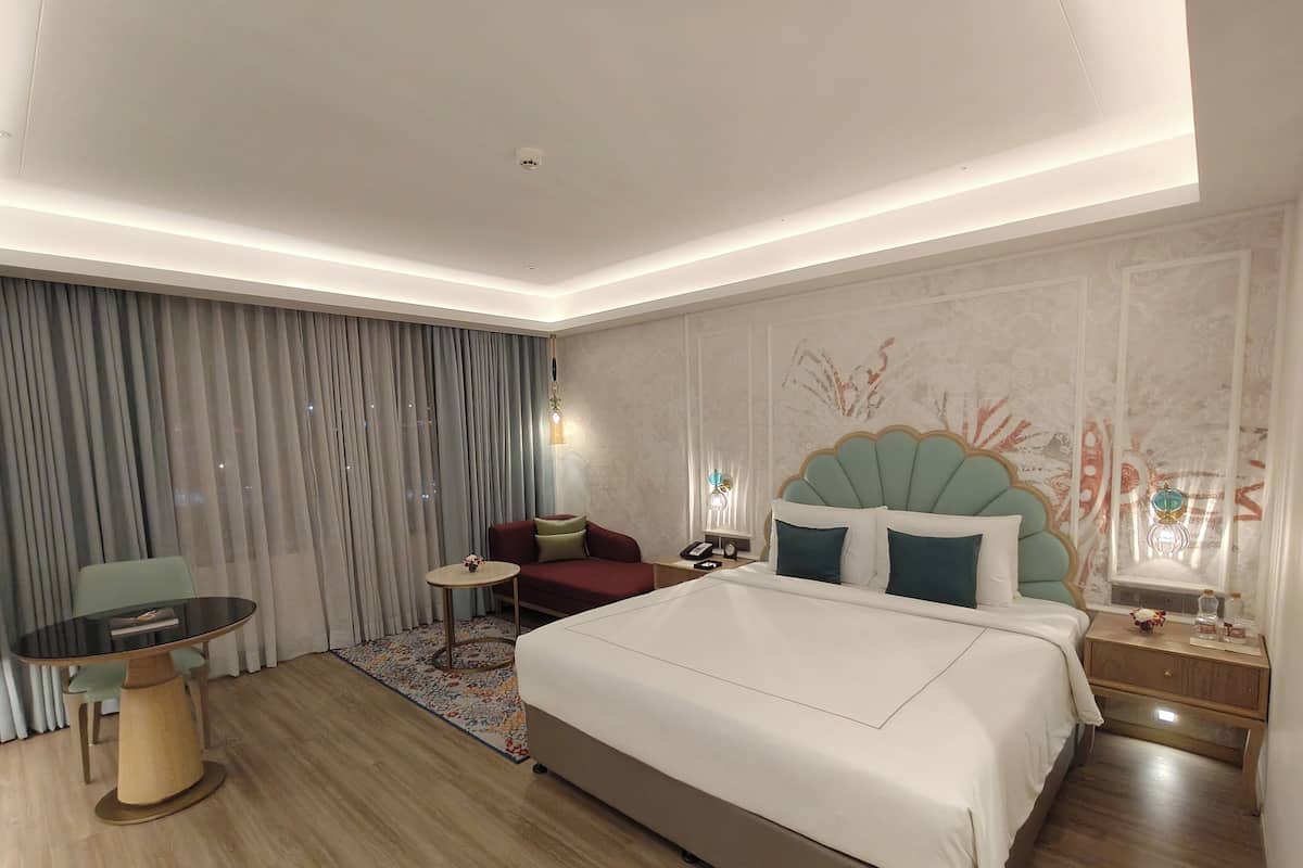A hotel room with a large bed, a sofa, a small round table, and a desk with a lamp.