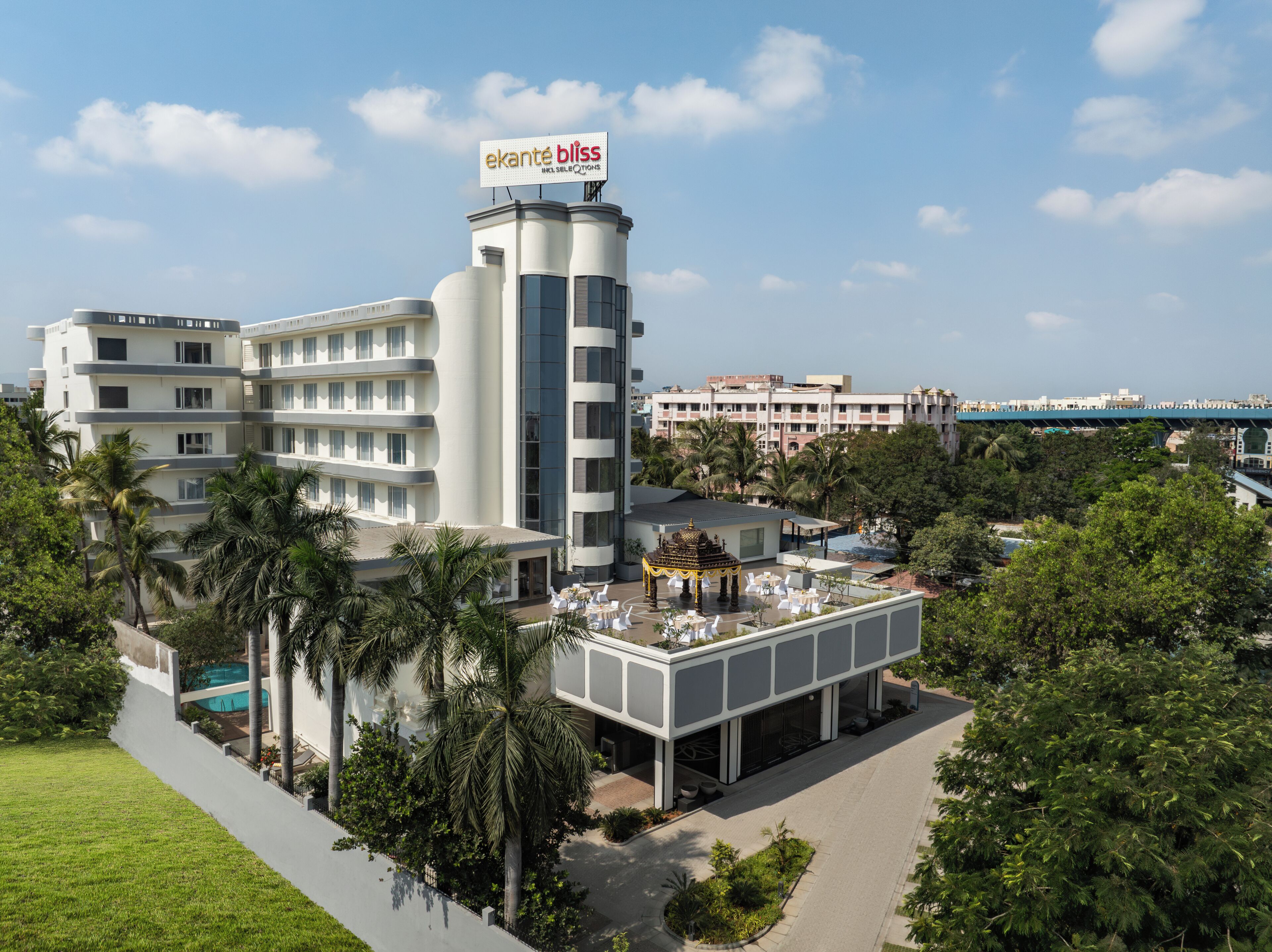 A modern hotel with a prominent sign, multiple floors, a rooftop pool area, and lush greenery.