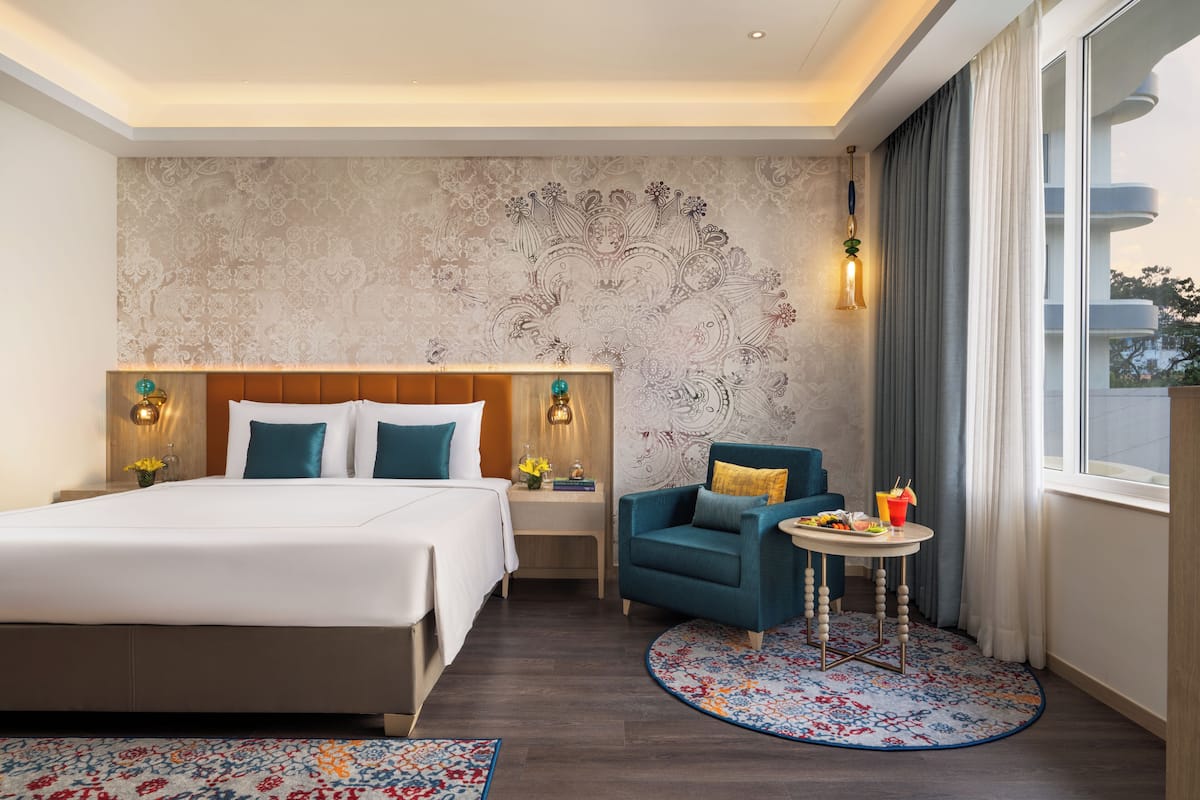 A hotel room with a large bed, a small table with a drink, a blue armchair, and a patterned wall.