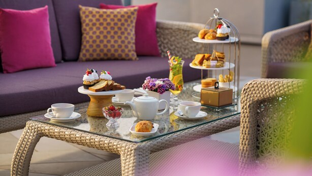 A glass-top table with a tea set, pastries, and a floral arrangement on an outdoor patio. - Ekante Bliss IHCL SeleQtions  (Tirupati)