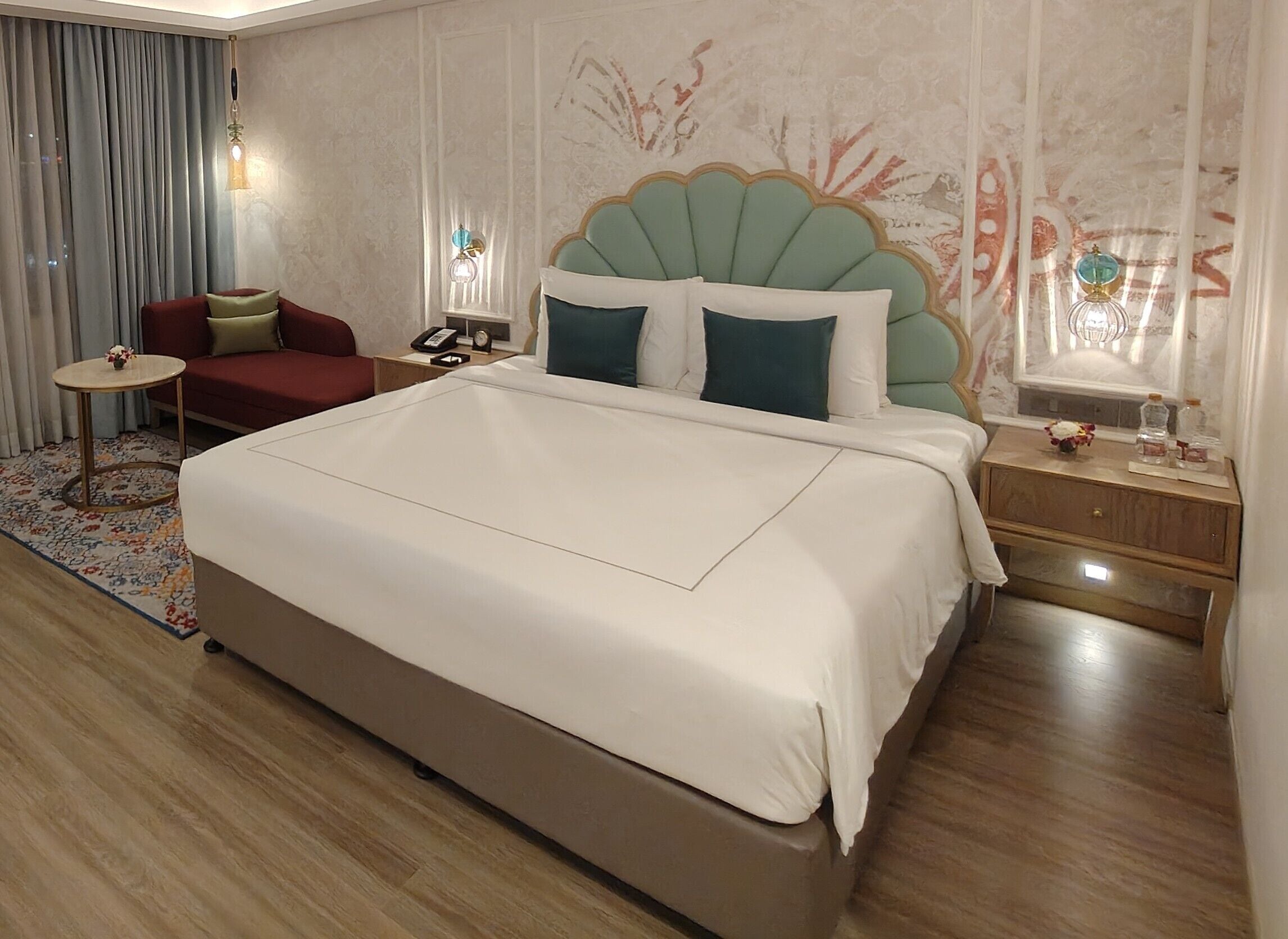 A large bed with a scalloped headboard, a bedside table with a lamp, a red armchair, and a small round table with a vase.