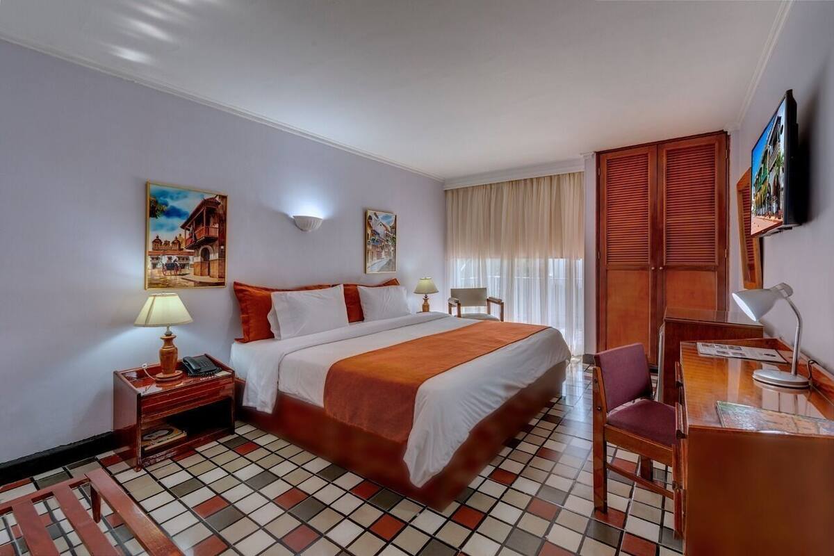 A hotel room with a bed, a desk, a chair, a TV, and artwork on the walls.