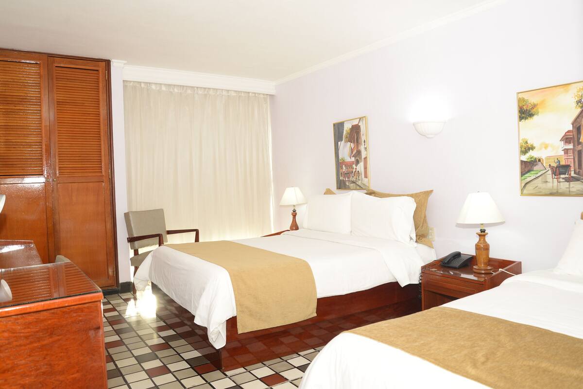 A hotel room with two beds, a desk, a chair, a nightstand with a lamp, and a window with curtains.