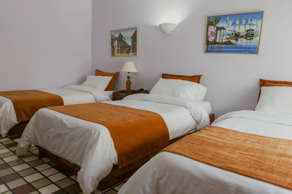 A hotel room with three beds, each with white bedding and orange throw blankets. There are two paintings on the walls, one depicting a street scene and the other a harbor with boats.