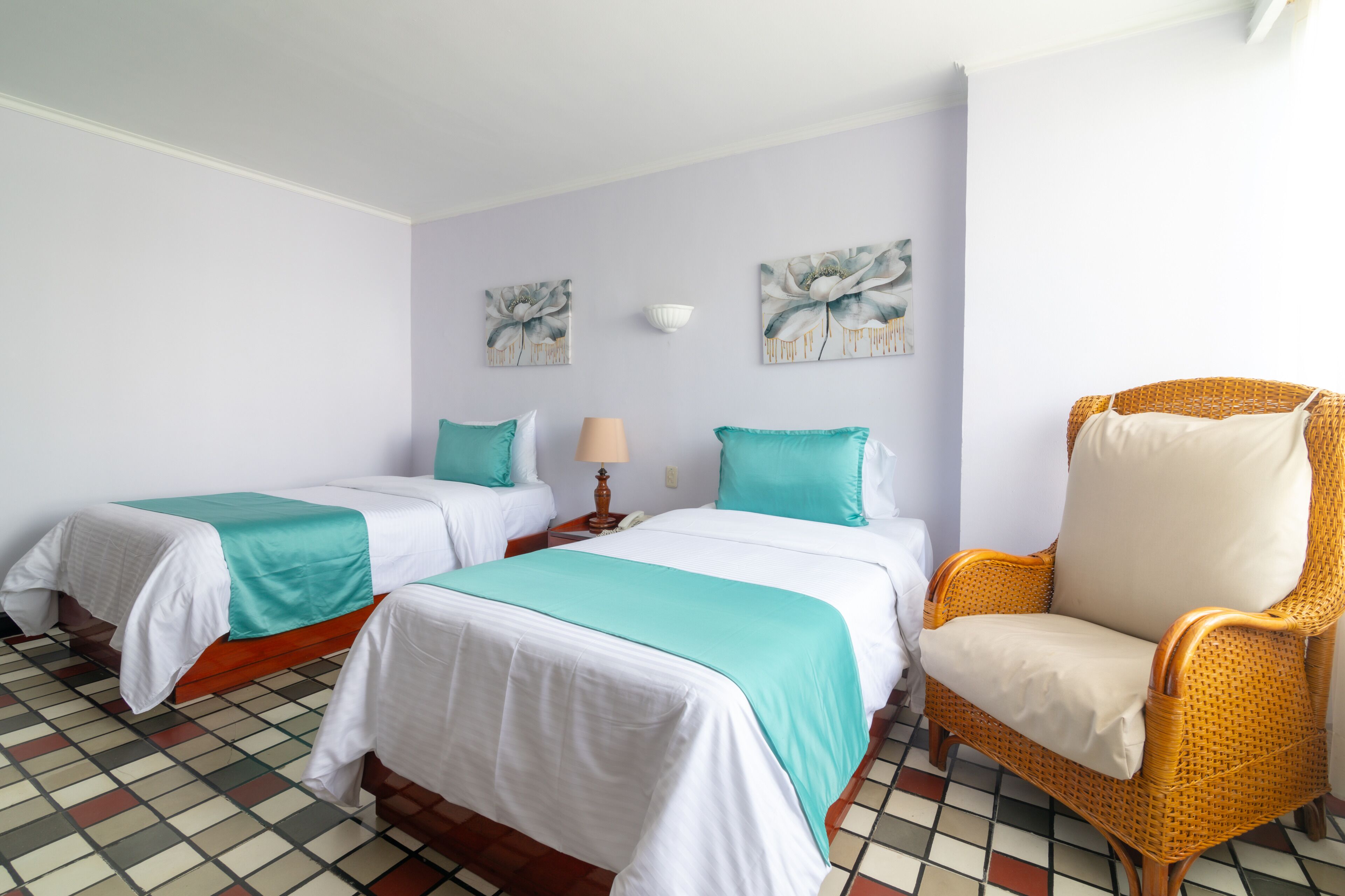 A hotel room with two beds, a wicker armchair, and a tiled floor.