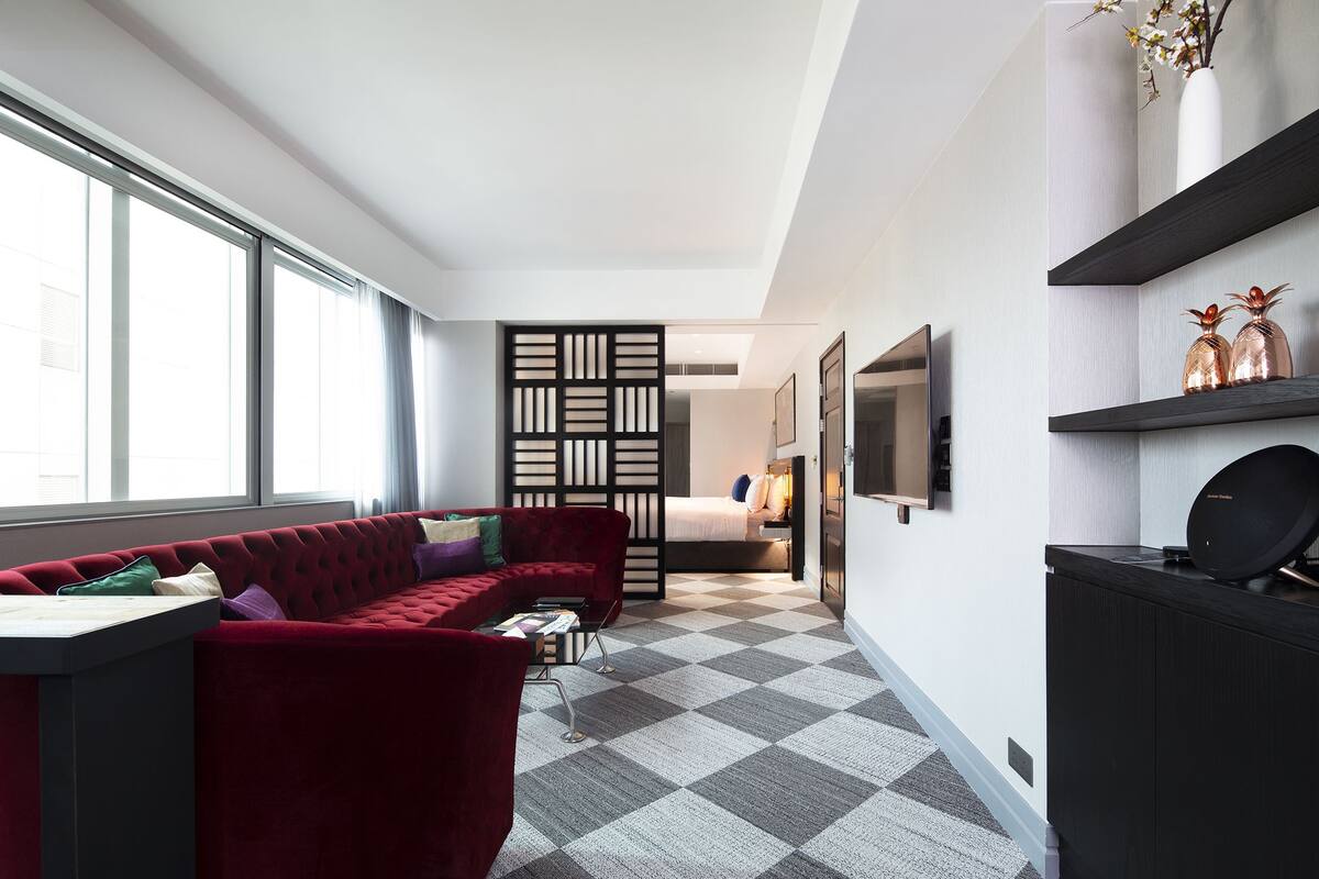 A modern hotel room with a red sofa, a desk, a large window, a black partition, a bed, a TV, and a decorative shelf.