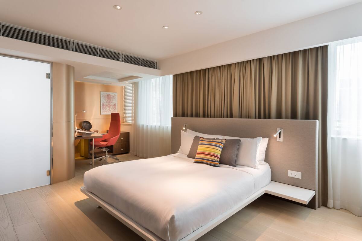 A modern hotel room with a large bed, a desk with a chair, and a view of the outdoors through the window.