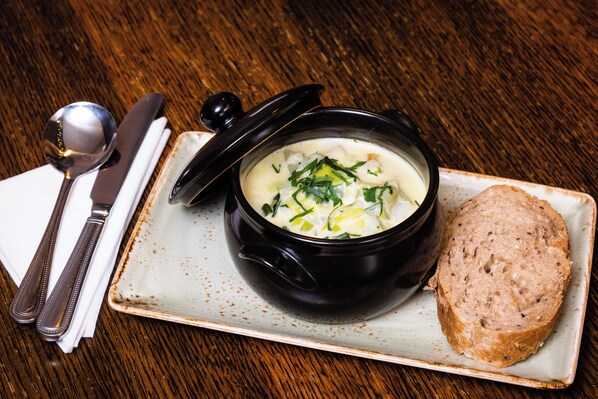 A bowl of soup with a lid on a plate, accompanied by a spoon and a knife on a napkin, and a slice of bread.