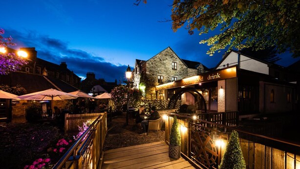 A wooden deck with a railing, a stone building with a sign, and outdoor seating with umbrellas. - The Old Mill Inn (Pitlochry)