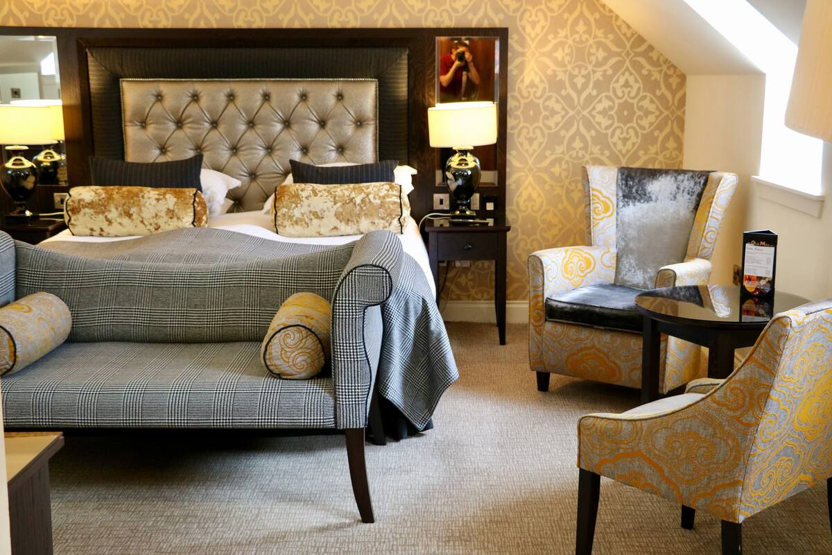 A hotel room with a large bed, a sofa, two armchairs, a nightstand with a lamp, and a mirror on the wall.