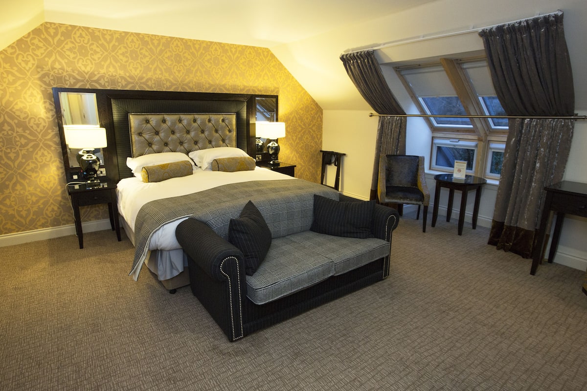 A hotel room with a large bed, a sofa, a chair, and a desk. There are two lamps on the nightstands, a mirror above the bed, and a window with curtains.