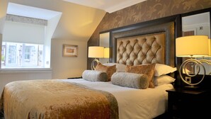 A hotel room with a large bed, two bedside lamps, a headboard with a tufted design, and a window with a view of buildings. - The Old Mill Inn (Pitlochry)